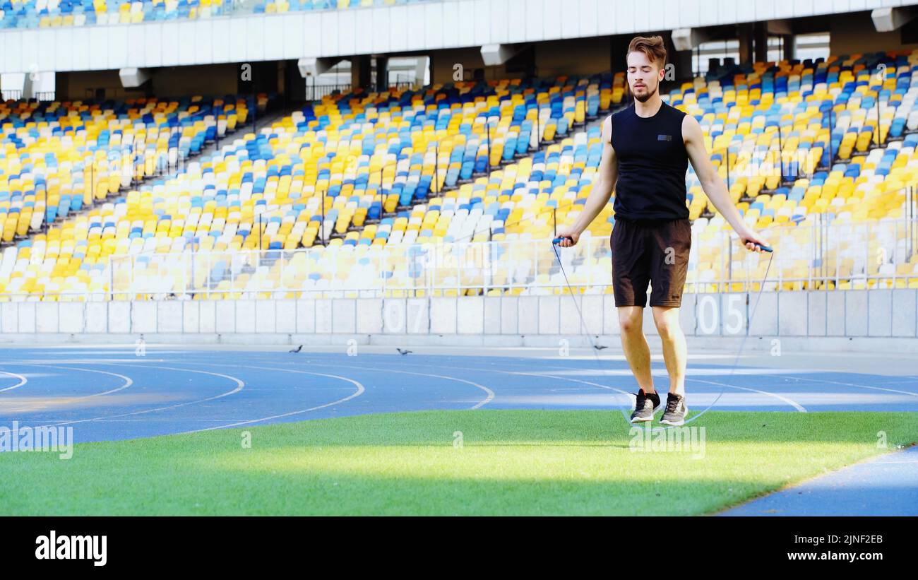 pleine longueur de sportif barbu sautant avec corde à sauter sur l'herbe verte au stade, image de stock Banque D'Images