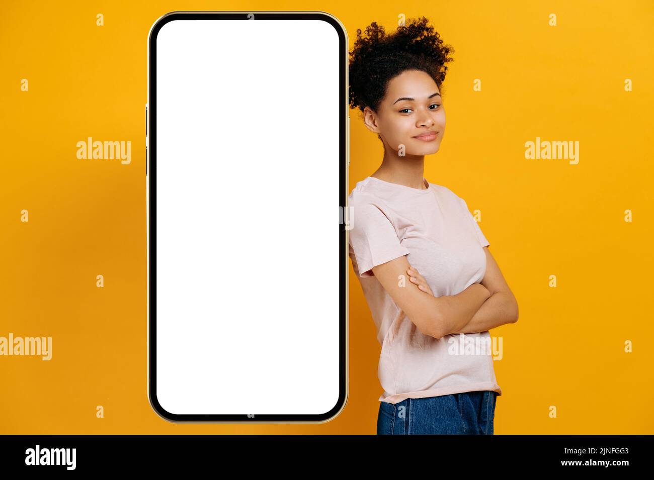 Jeune afro-américaine positive et curieuse, se tient près d'un smartphone agrandi avec un écran maquette blanc pour la présentation ou la publicité, regarde la caméra, sourit, arrière-plan isolé orange Banque D'Images