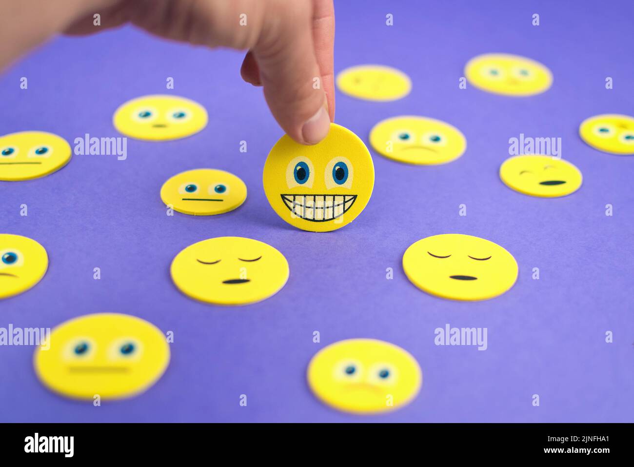 Gros plan de la main tenant un smiley jaune visage emoji entre les émoticônes tristes sur fond violet Banque D'Images