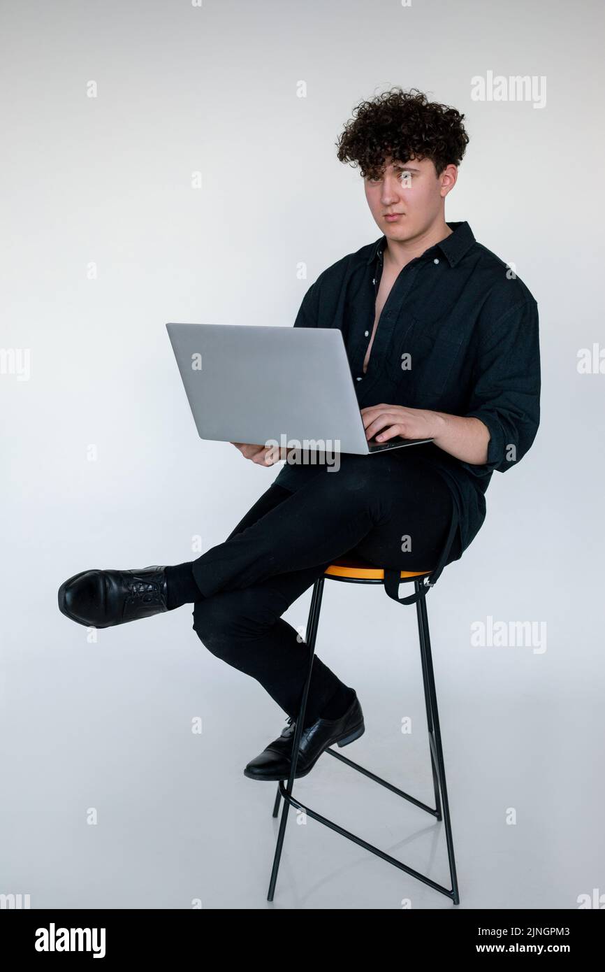 Photo pleine longueur de jeune homme fatigué en forme de curly dans l'aspect noir total assis en croix legged sur la chaise, dactylographiant et travaillant sur un ordinateur portable moderne, studio gris Banque D'Images