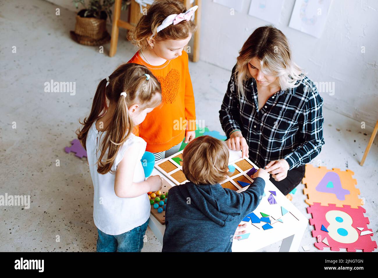 Vue de dessus de groupe d'enfants d'âge préscolaire et d'enseignant. Jeune femme montrant comment jouer à des jeux de société collectant des figures géométriques. Banque D'Images