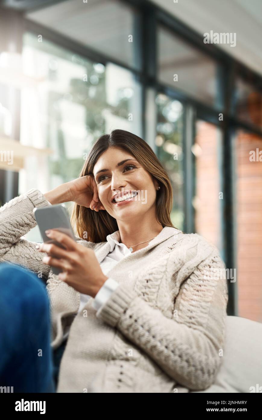 Peut-être devrais-je reconsidérer mon point de vue : une jeune femme attirante qui a l'air pensive et qui envoie des SMS tout en se relaxant sur son canapé à la maison. Banque D'Images