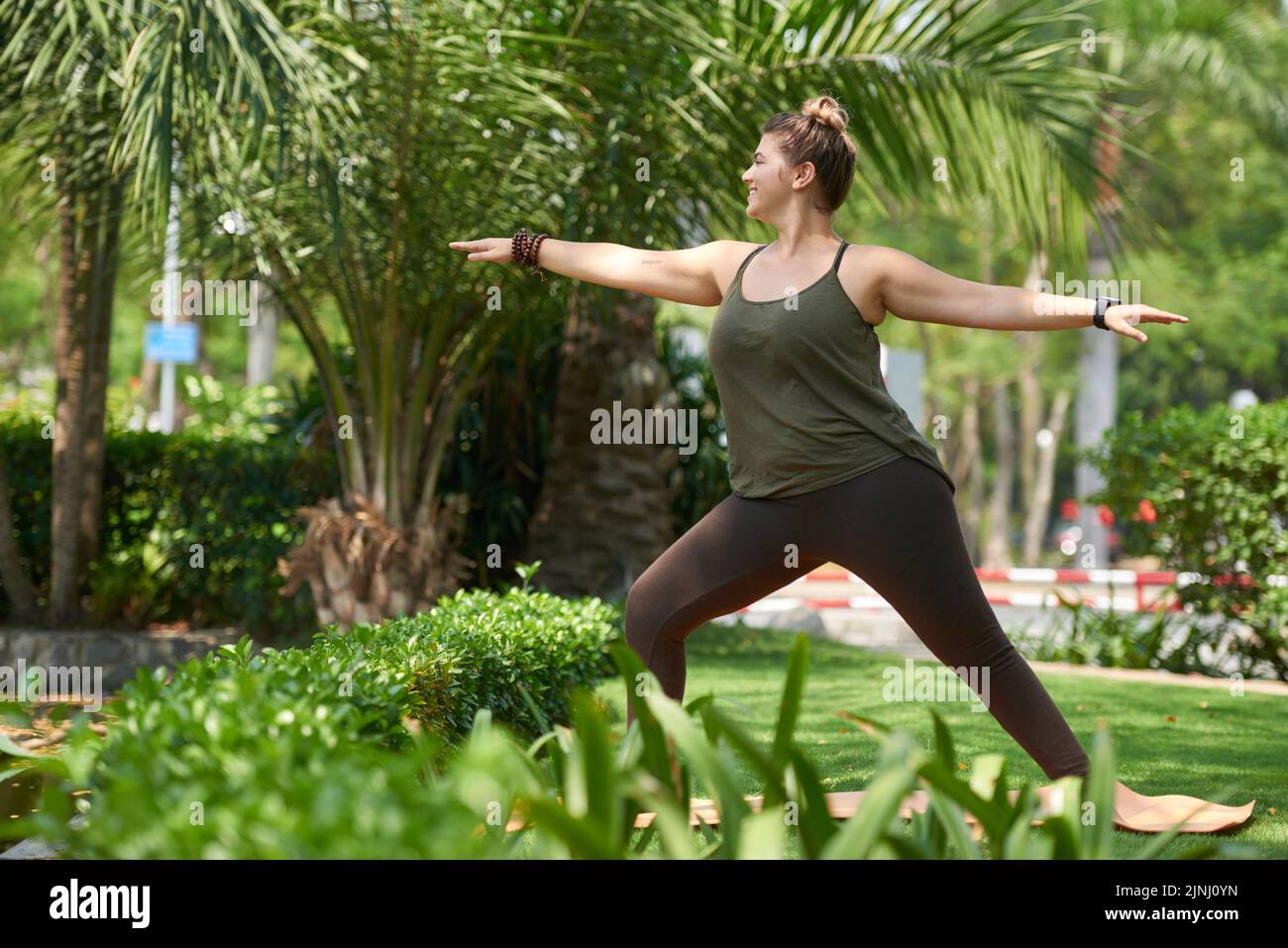 Belle femme obèse portant dessus et leggings debout sur le tapis de yoga et faisant l'exercice d'étirement tout en ayant l'entraînement à l'extérieur Banque D'Images