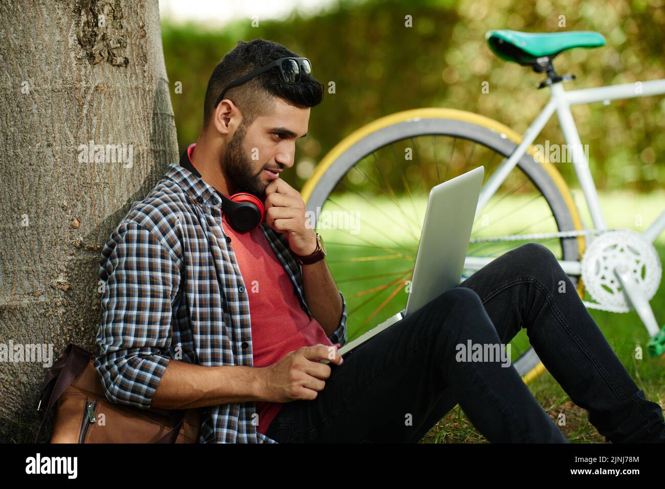 Beau jeune étudiant assis sur la pelouse du parc et utilisant un ordinateur portable moderne tout en se préparant à l'examen universitaire, vélo moderne debout derrière lui Banque D'Images