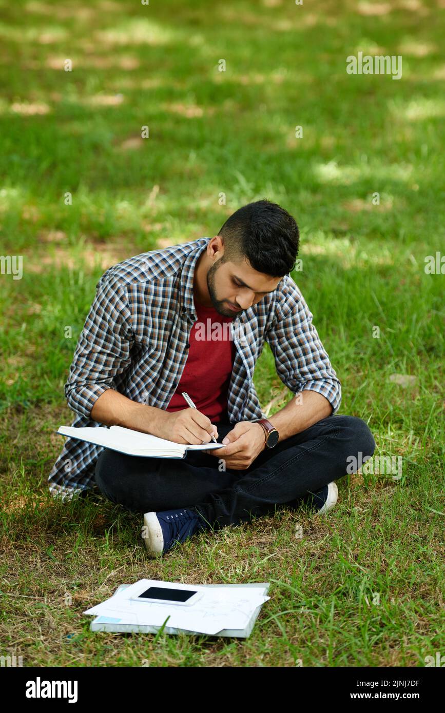 Étudiant indien concentré en vêtements décontractés assis en position lotus sur la pelouse verte et prenant des notes tout en préparant l'examen universitaire Banque D'Images