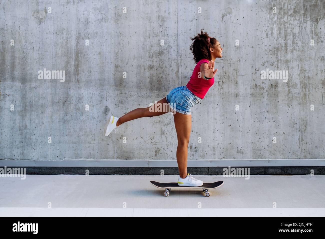 Adolescente multiraciale à cheval sur un skateboard devant un mur en béton, debout dans une jambe, en équilibre. Vue latérale. Banque D'Images