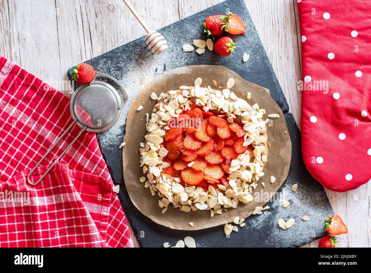 Gâteau à la galette maison avec fraises et amandes, placé sur un bureau noir et une table en bois. Serviette rouge avec outils de cuisson sur le côté Banque D'Images