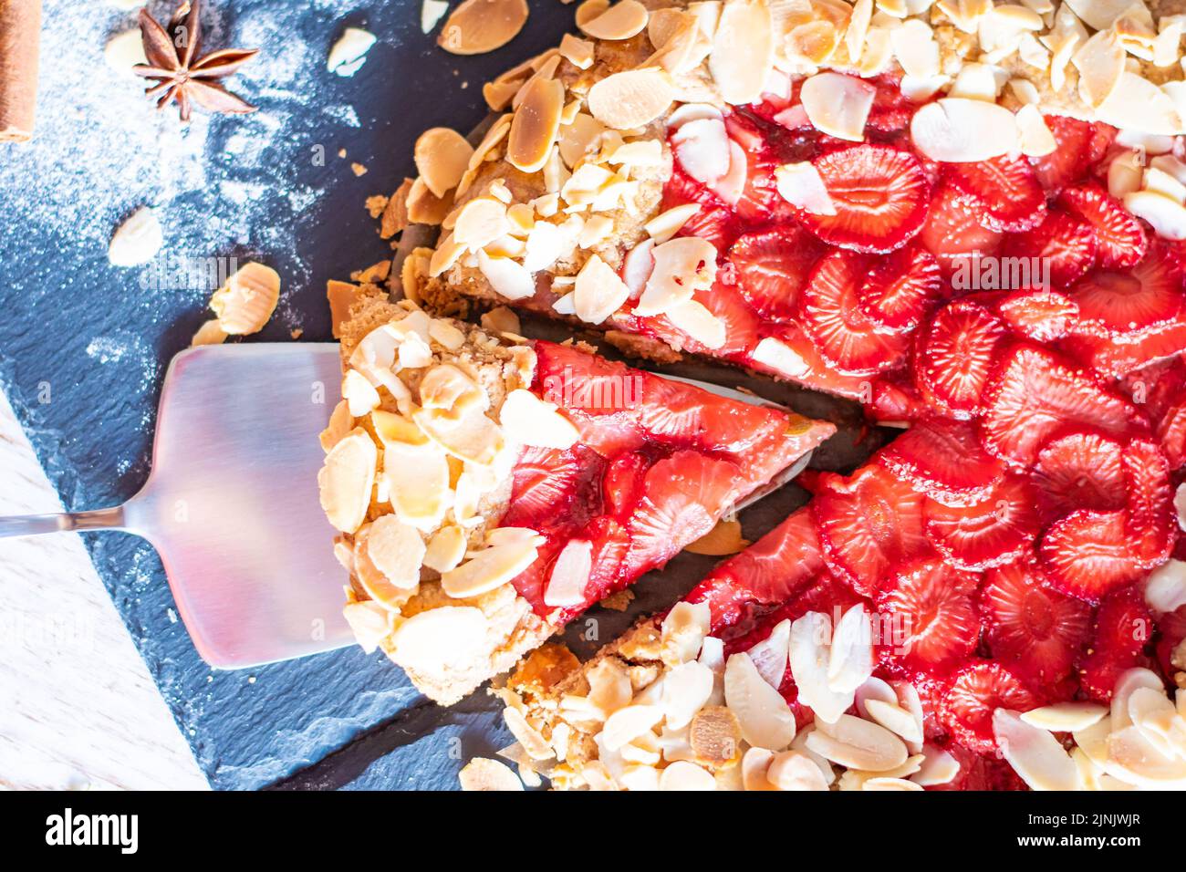 Gâteau à la galette maison avec fraises et amandes, placé sur un bureau noir et une table en bois. Serviette rouge avec outils de cuisson sur le côté Banque D'Images