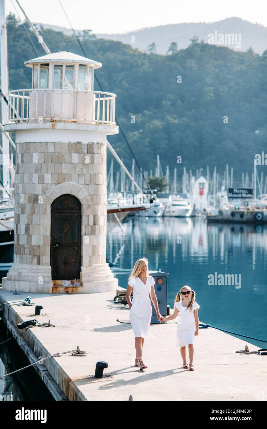 Bonne mère millénaire et fille blonde adolescente vêque de robes blanches marchant au bord du port et de la marina avec des yachts dans une station balnéaire touristique avec voile Banque D'Images