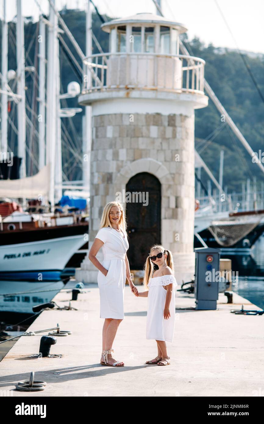 Bonne mère millénaire et fille blonde adolescente vêque de robes blanches marchant au bord du port et de la marina avec des yachts dans une station balnéaire touristique avec voile Banque D'Images