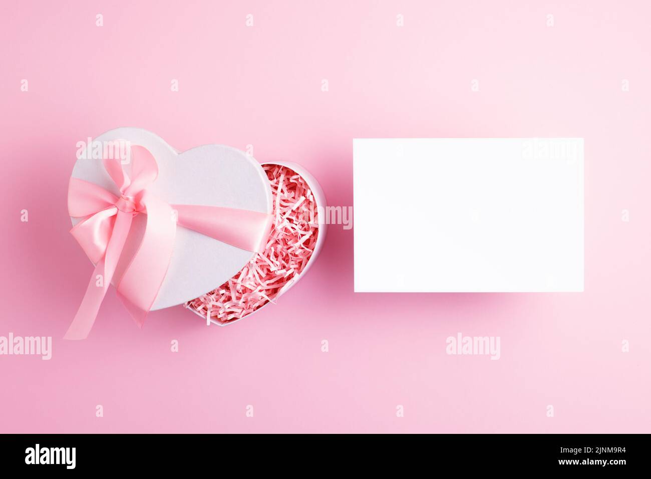 Publicité, maquette promotionnelle de vacances avec carte vide et magnifique boîte cadeau en forme de coeur de fête sur fond rose clair. Vue de dessus, espace de copie. Banque D'Images
