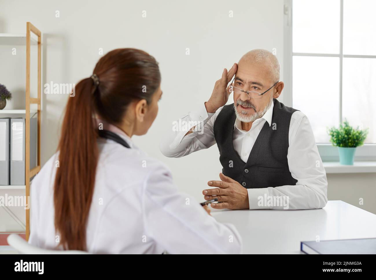 Un homme plus âgé lors d'une visite ou d'une consultation à la clinique informe le médecin de ses maux de tête graves. Banque D'Images