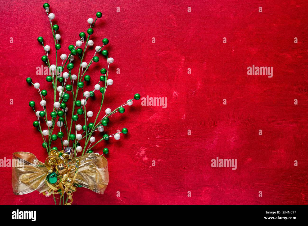 thème des fêtes avec des branches ornées de perles vertes et blanches attachées dans un noeud en or métallique et des cloches de jingle sur un fond rouge texturé Banque D'Images