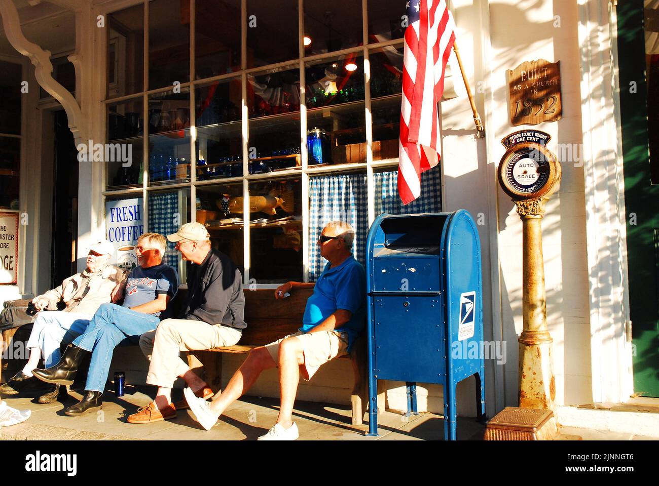 Un groupe d'amis seniors s'engage dans la conversation et la discussion assis sur un banc à l'extérieur d'un magasin général lors d'une journée ensoleillée de vacances d'été Banque D'Images