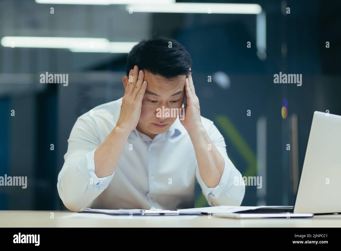 Homme d'affaires stressé tenant le visage avec ses mains parce qu'il a des problèmes. L'homme asiatique en faillite est assis avec un ordinateur portable et des documents sur le bureau au lieu de travail dans le bureau. Erreur d'affaires financière. Banque D'Images