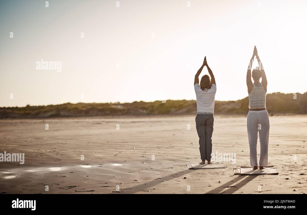 La plage est tranquille, ajoutez du yoga et elle devient libératrice. Photo d'un couple méconnaissable pratiquant le yoga sur la plage. Banque D'Images