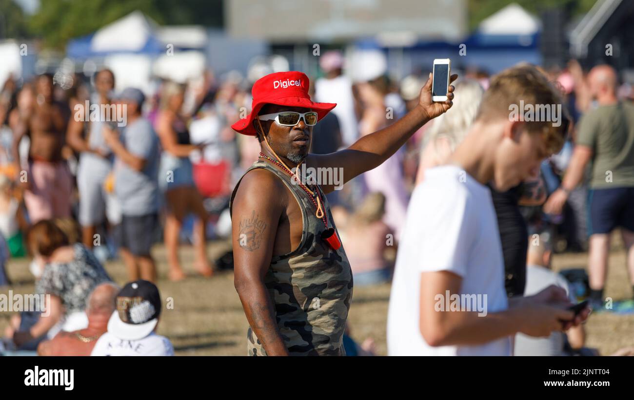 Un homme noir avec un chapeau rouge et des lunettes de soleil blanches tient un téléphone cellulaire dans les airs lors d'un festival en plein air Banque D'Images