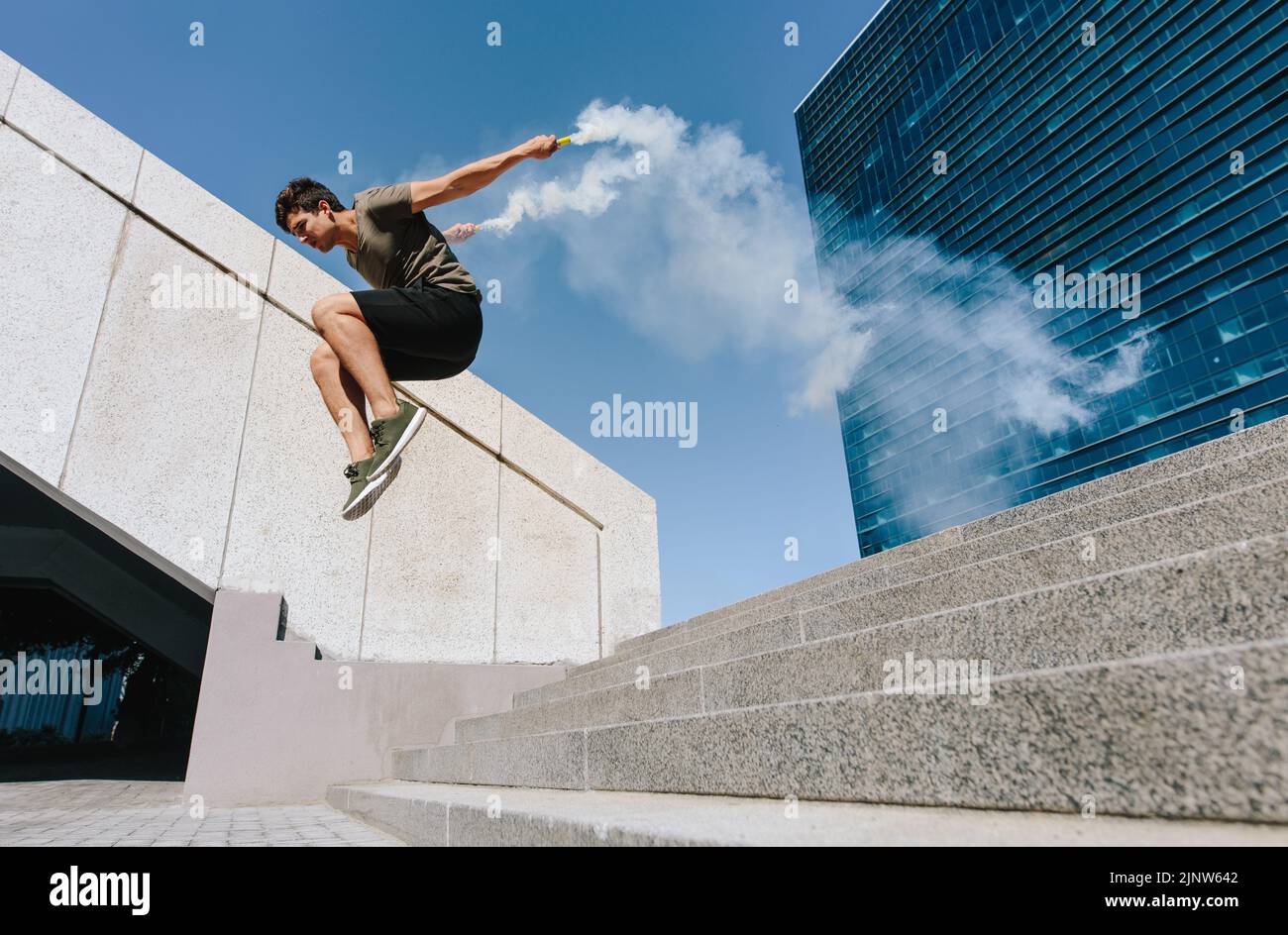 Jeune homme sautant à l'extérieur avec des grenades de fumée. Chemin libre sautant sur quelques marches dans un espace urbain. Banque D'Images