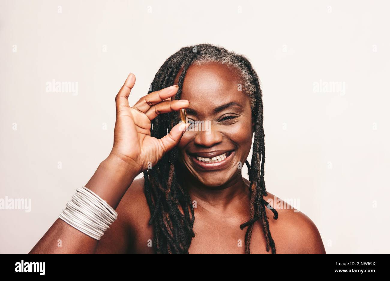 Gaie femme mûre souriant à la caméra tout en tenant une capsule d'acides gras oméga 3. Bonne femme avec des dreadlocks en utilisant des suppléments nutritionnels Banque D'Images