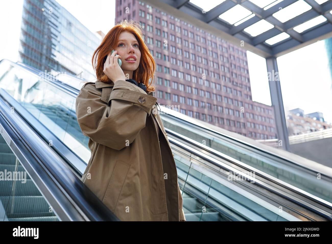 Une jeune fille à tête rouge parle sur un téléphone portable, debout sur un escalier roulant. Banque D'Images