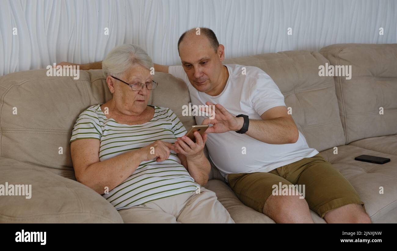 Dans la salle de séjour d'un côté du canapé, assis une vieille grand-mère de 80s ans avec un adulte de 40s fils, une femme tenant un smartphone montrant à l'homme plus âgé nouvelle application, teac Banque D'Images