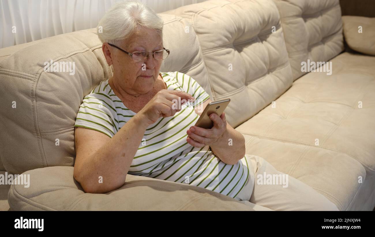 Une femme âgée agréable reposant sur un canapé, utilisant des applications sur un smartphone. Bonne vieille femme mûre discutant dans la messagerie ou le réseau social avec ven Banque D'Images