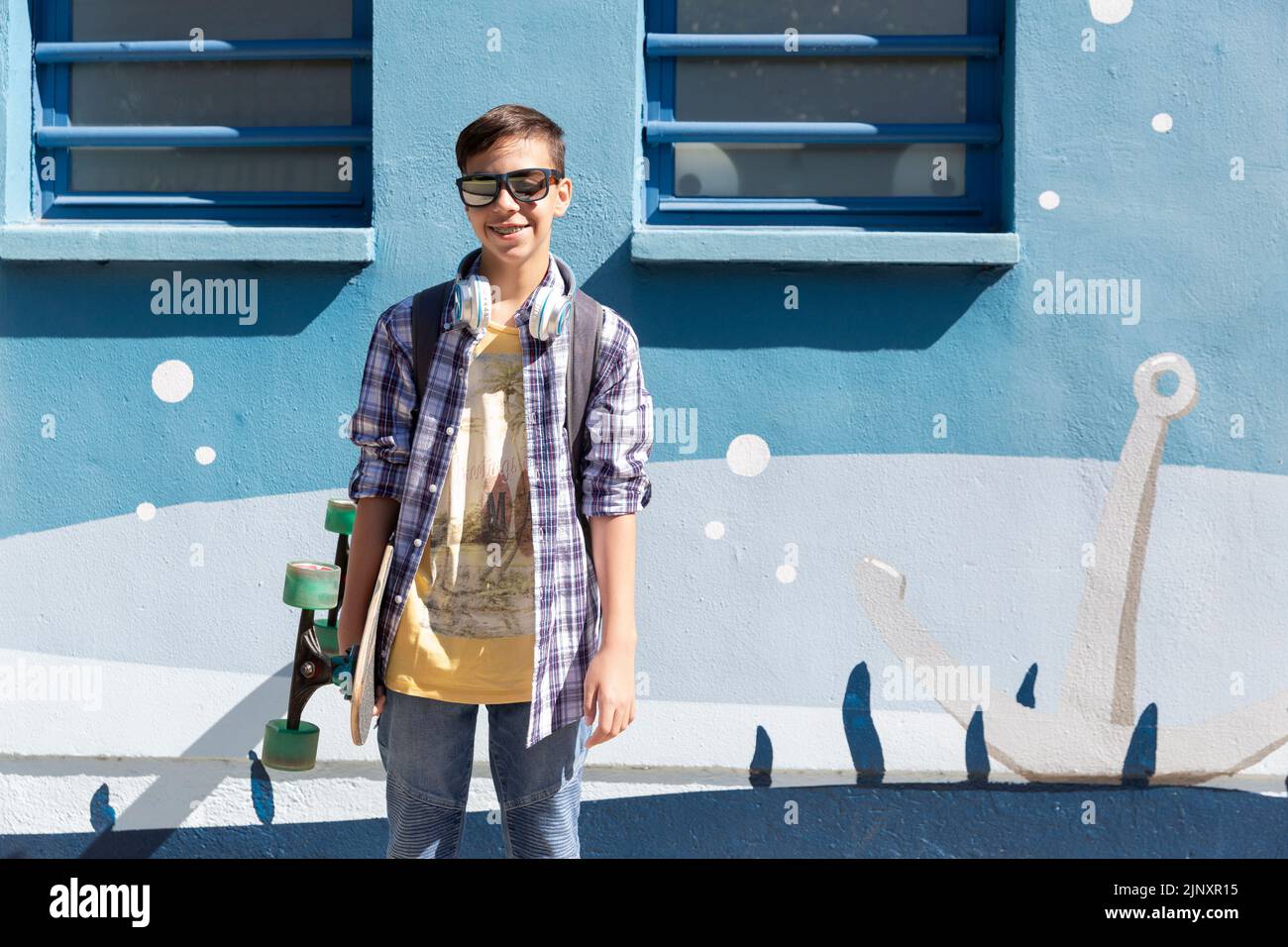 Jeune garçon caucasien souriant regardant la caméra avec son skateboard à côté d'un mur bleu. Banque D'Images