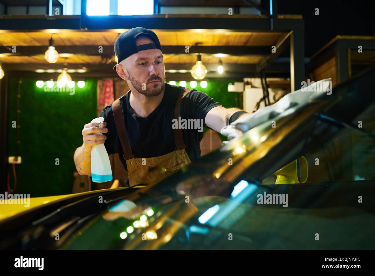 Jeune homme de service d'entretien tenant une bouteille en plastique avec pulvérisateur et essuyant le pare-brise avec plumeau tout en le nettoyant Banque D'Images