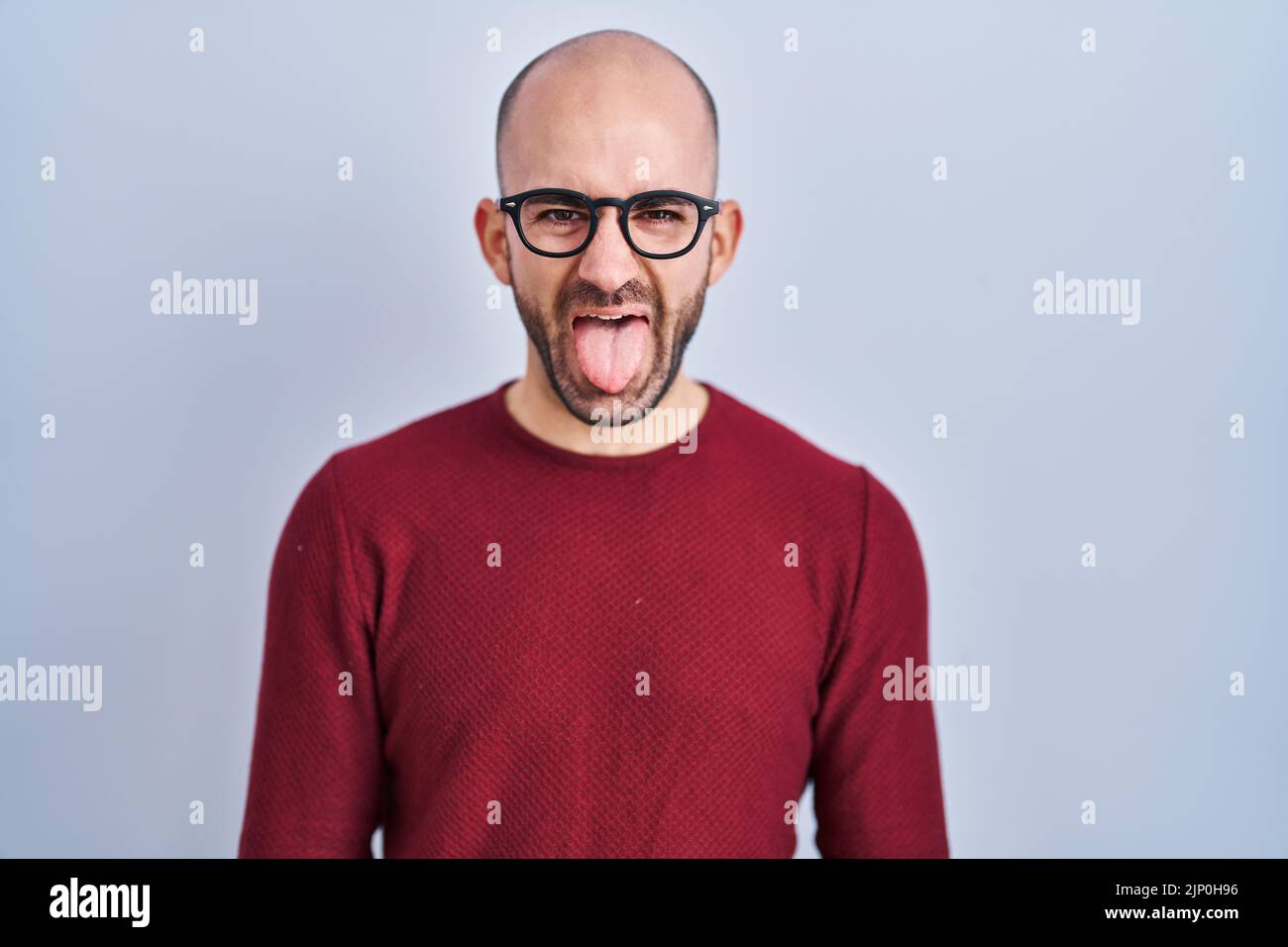 Jeune homme chauve avec barbe debout sur fond blanc portant des lunettes collant la langue heureux avec l'expression drôle. Concept d'émotion. Banque D'Images