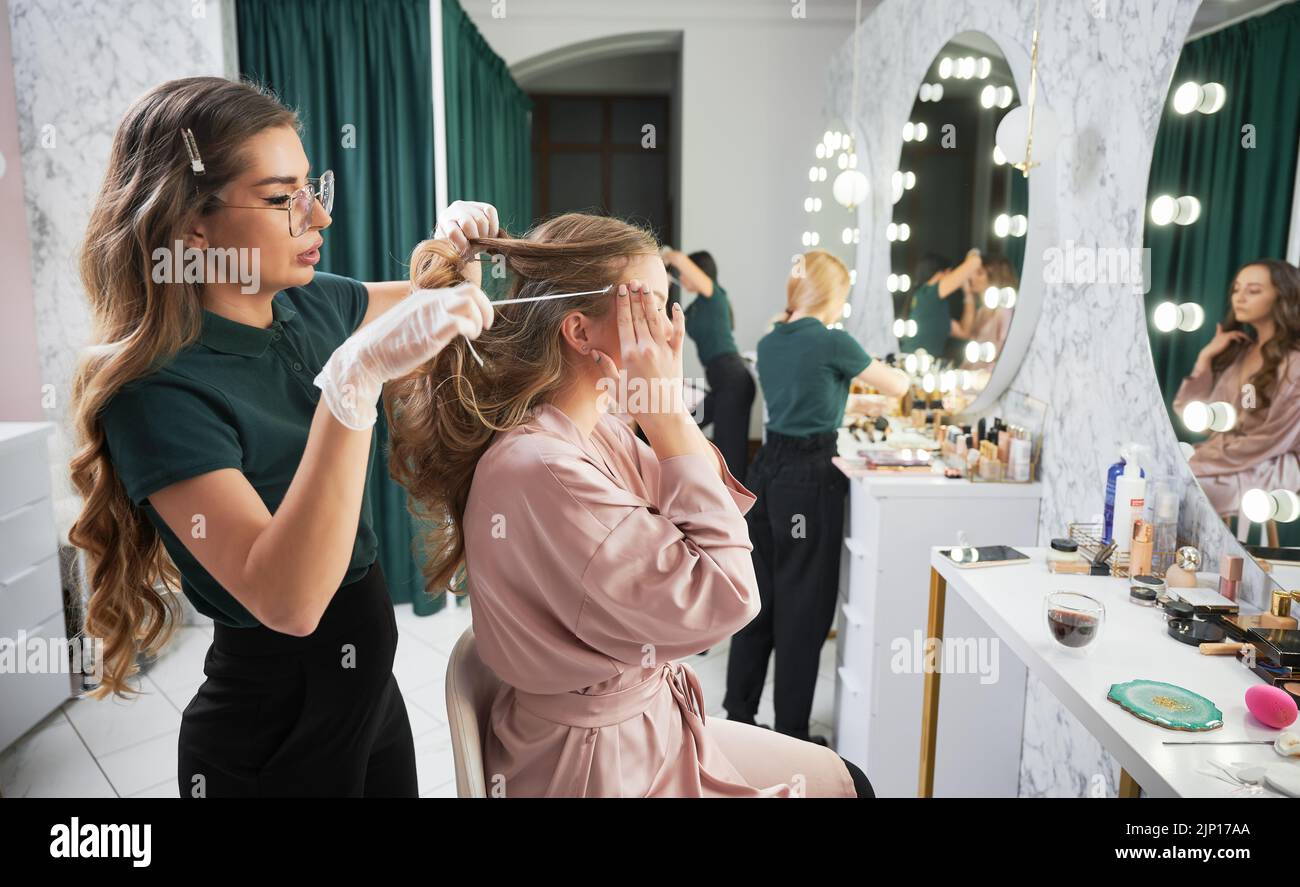 Femme assise à la coiffeuse et touchant le temple tout en ayant la procédure de beauté en salon. Styliste féminine avec des gants stériles tenant la mèche de cheveux du client et tirant la corde tout en travaillant dans le studio de visualisation. Banque D'Images