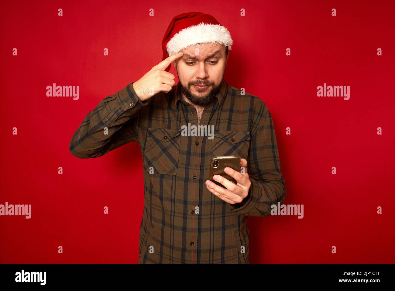 un homme sérieux réfléchis en chapeau de noël lit sur le téléphone tient l'index à sourcil essaie de comprendre Banque D'Images