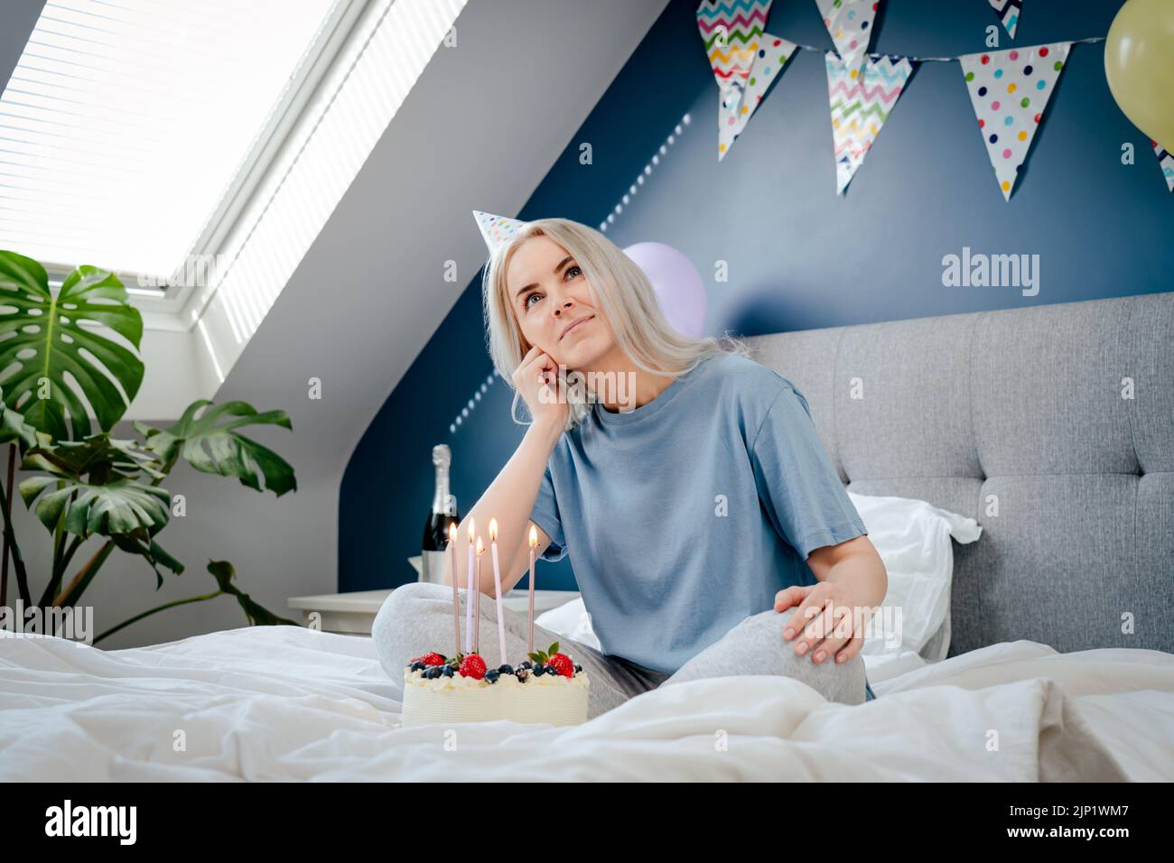 Femme souriante et attentive dans un chapeau de fête d'anniversaire avec gâteau de fête et bougies sur le lit dans la chambre décorée. Faites un souhait. Surprise du matin. Banque D'Images Femme souriante et attentive dans un chapeau de fête d'anniversaire avec gâteau de fête et bougies sur le lit dans la chambre décorée. Faites un souhait. Surprise du matin. Banque D'Images
