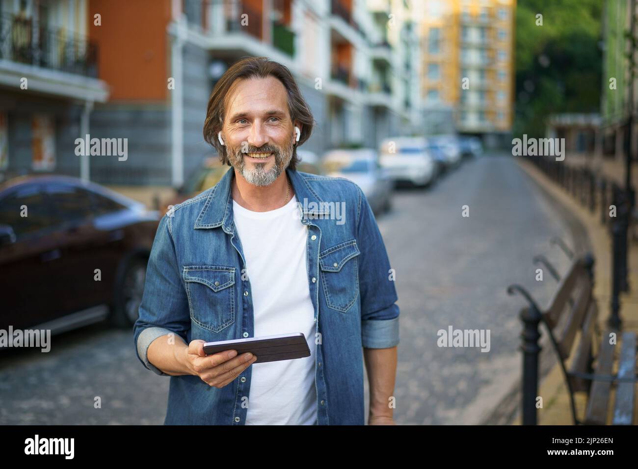 Un travailleur indépendant gris d'âge moyen tient une tablette numérique dans les mains pour passer un appel tout en restant dans les rues urbaines. Homme mature parlant ou écoutant de la musique utilisez des écouteurs sans fil pour voyager dans la vieille ville. Banque D'Images
