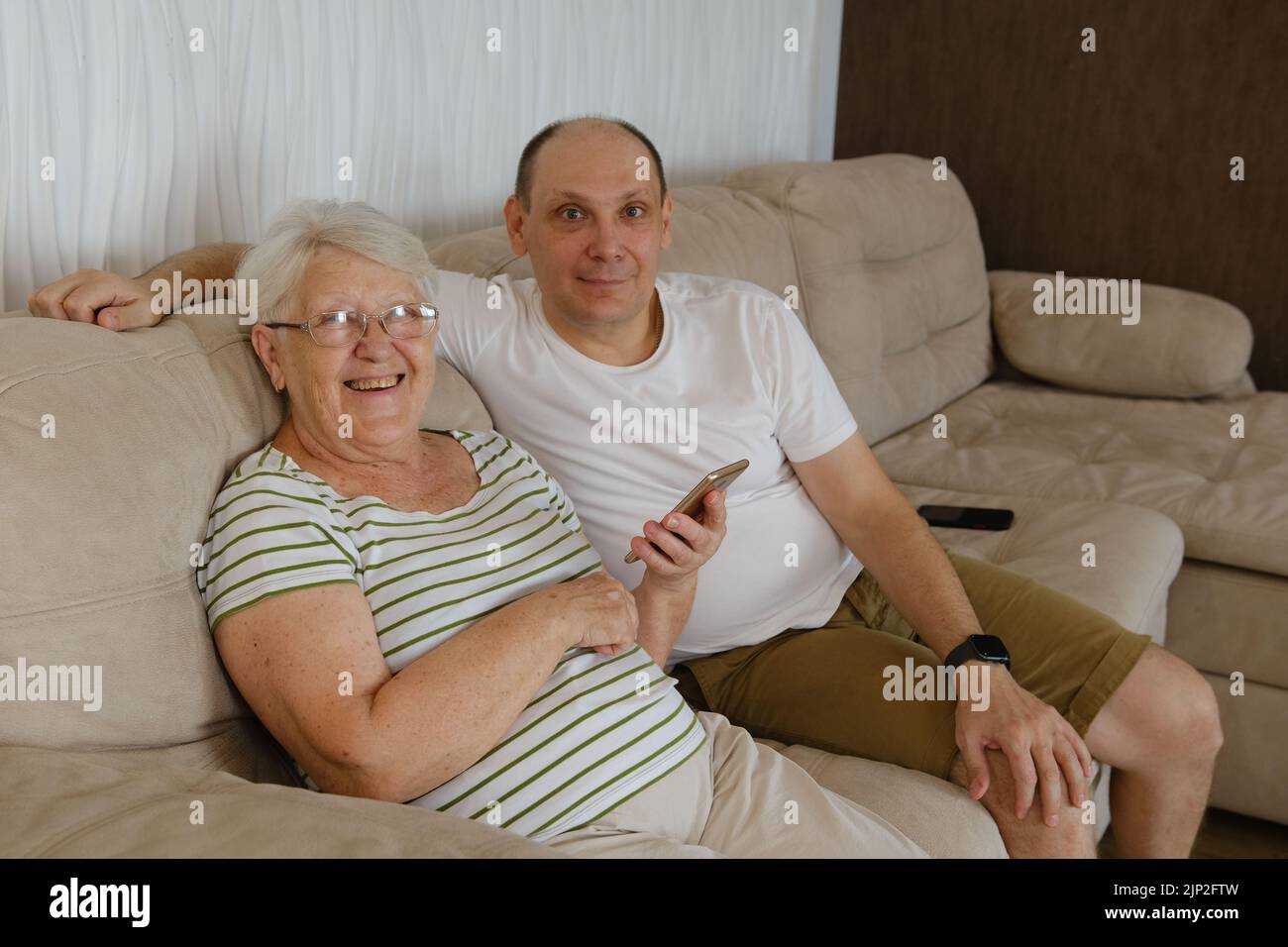 Dans la salle de séjour d'un côté du canapé, assis une vieille grand-mère de 80s ans avec un adulte de 40s fils, une femme tenant un smartphone montrant à l'homme plus âgé nouvelle application, teac Banque D'Images