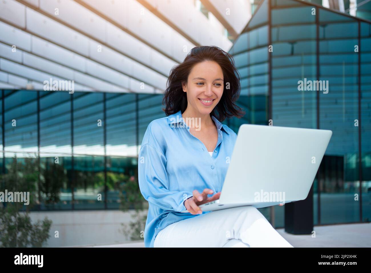 Jeune femme d'affaires souriante utilisant un ordinateur portable tout en étant assise à l'extérieur du bâtiment de bureau. Femme d'affaires confiante travaillant avec un ordinateur portable dans le quartier financier. Banque D'Images