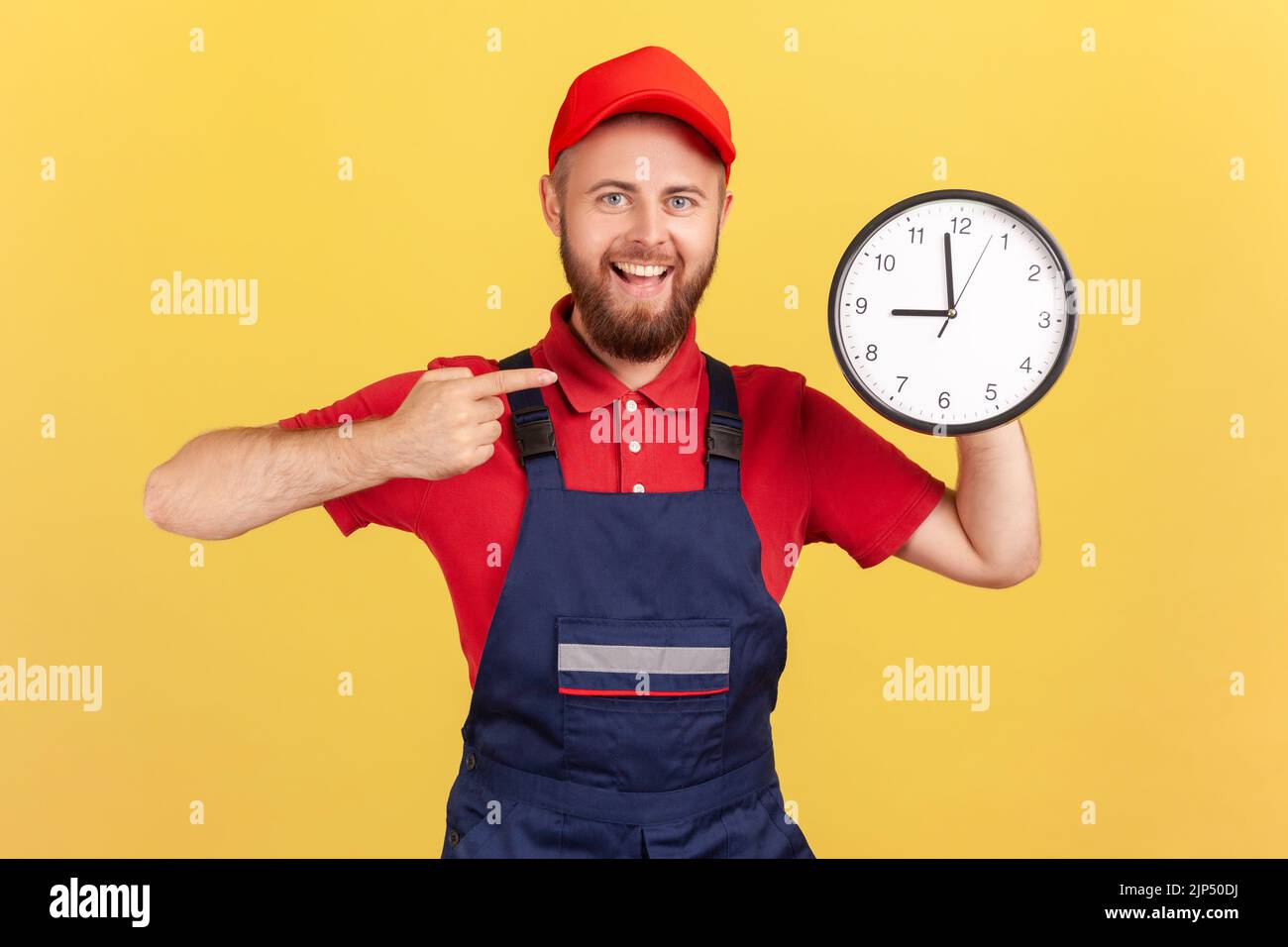 Portrait de l'homme ravi travailleur debout et pointant vers la grande horloge murale dans ses mains, regardant l'appareil photo, portant une combinaison et une casquette rouge. Studio d'intérieur isolé sur fond jaune. Banque D'Images