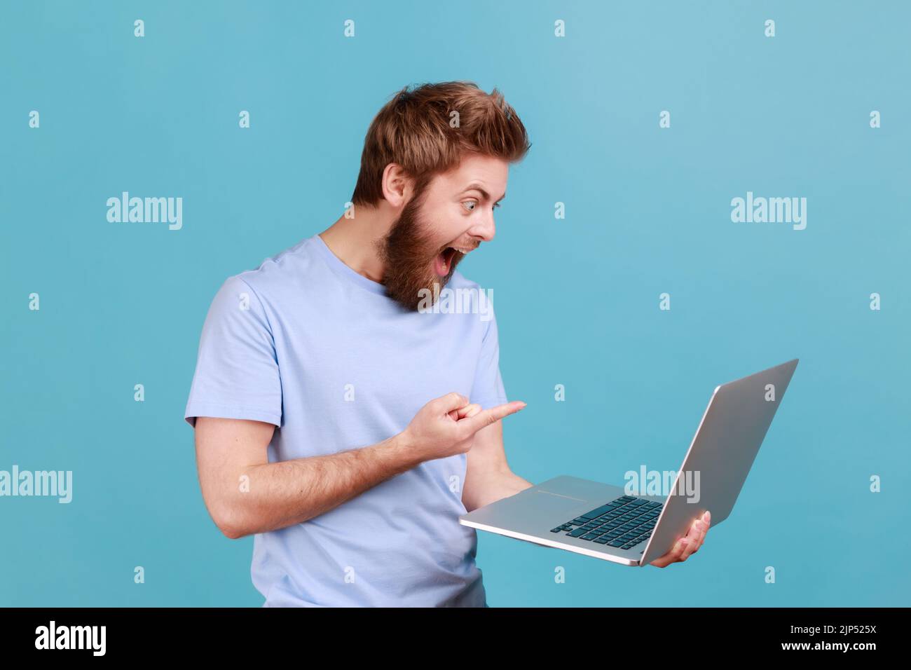 Portrait d'un beau homme surpris surpris barbu pointant le doigt sur l'écran d'un ordinateur portable avec de grands yeux choqués par le contenu. Studio d'intérieur isolé sur fond bleu. Banque D'Images