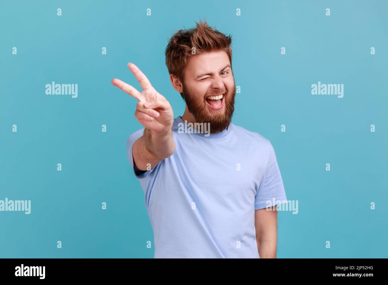 Portrait de l'homme heureux optimiste barbu faisant le geste de la victoire et wencing playentilly à l'appareil photo, montrant la paix, le signe v avec les deux doigts. Studio d'intérieur isolé sur fond bleu. Banque D'Images