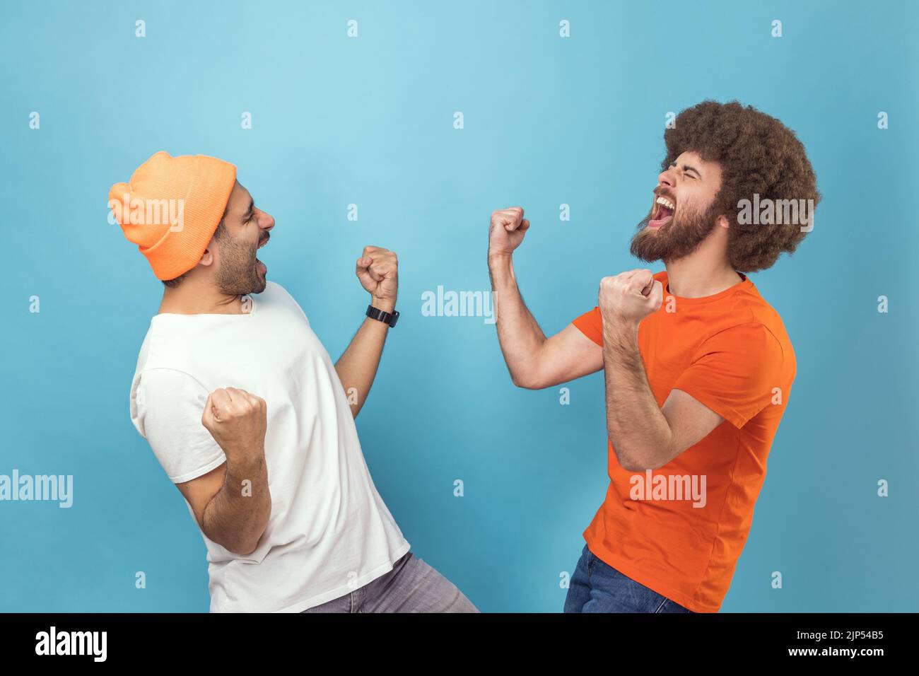 Portrait de deux jeunes adultes hipster hommes montrant oui geste et criant célébrant la victoire, le succès, les rêves deviennent vrai, l'euphorie. Studio d'intérieur isolé sur fond bleu. Banque D'Images Portrait de deux jeunes adultes hipster hommes montrant oui geste et criant célébrant la victoire, le succès, les rêves deviennent vrai, l'euphorie. Studio d'intérieur isolé sur fond bleu. Banque D'Images