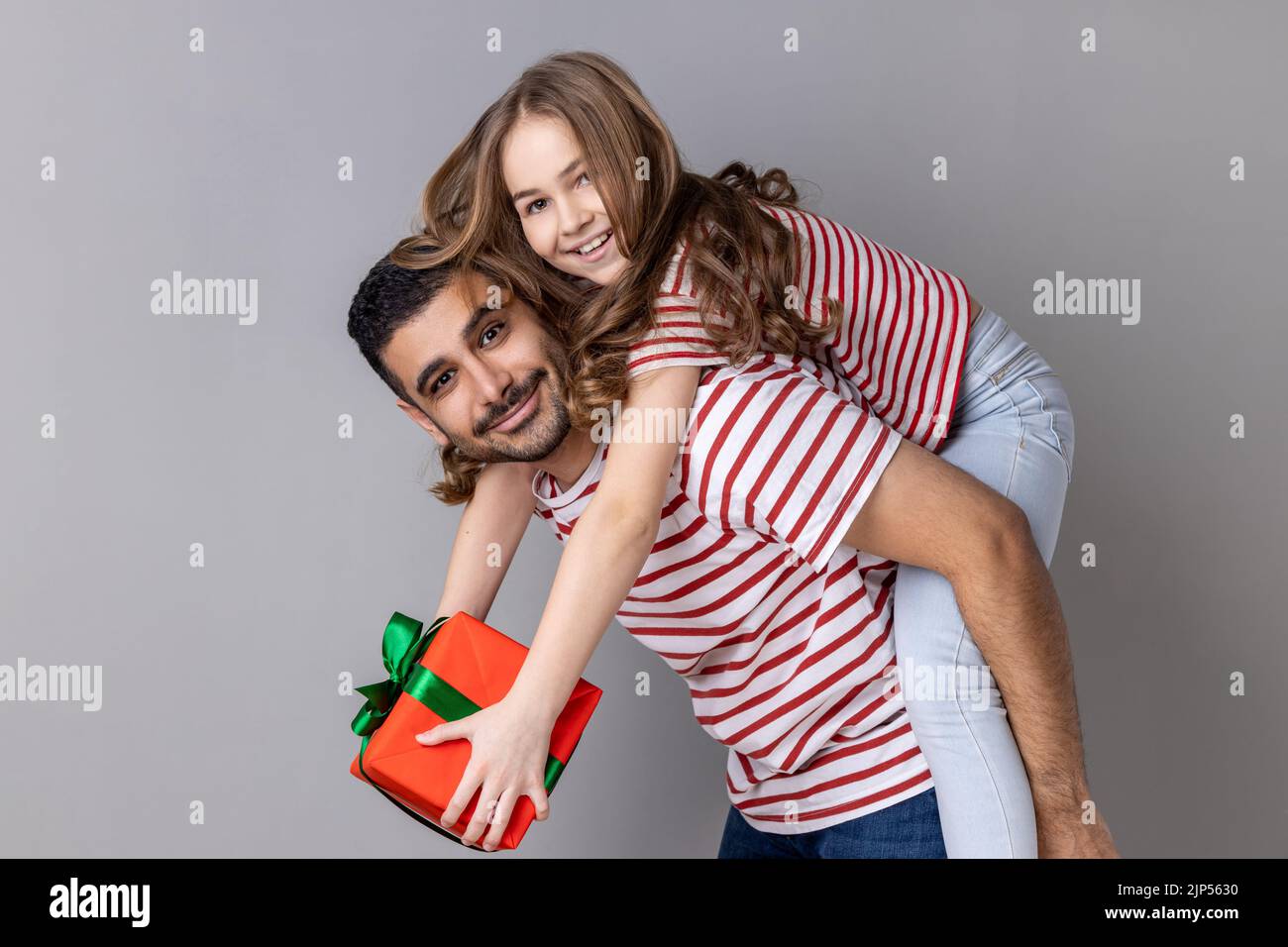 Portrait d'un père et d'une fille extrêmement heureux en T-shirts rayés célébrant ensemble les vacances, papa tenant un enfant avec une boîte cadeau sur son dos. Prise de vue en studio isolée sur fond gris. Banque D'Images