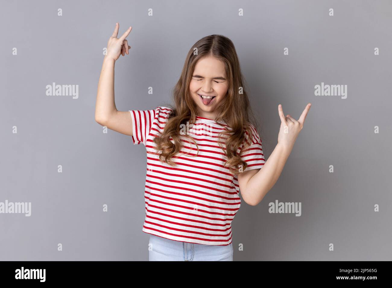 Portrait d'une petite fille portant un T-shirt rayé montrant rock and roll geste Heavy Metal signe, aime la musique préférée, a du plaisir, exclame de joie. Prise de vue en studio isolée sur fond gris. Banque D'Images