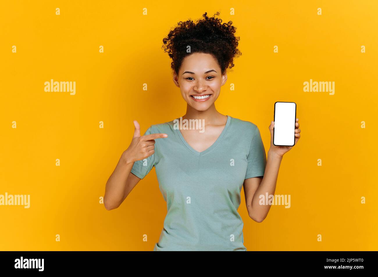 Écran de maquette. Positive african american jeune femme, regarde la caméra heureusement, tient le téléphone cellulaire avec écran blanc vide et pointe le doigt dessus, se tient sur un arrière-plan isolé orange, souriant Banque D'Images