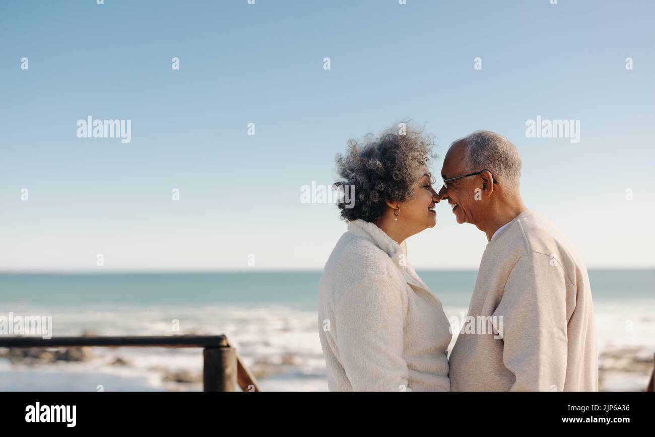 Être romantique au bord de l'océan. Heureux couple senior souriant et touchant leurs nez ensemble tout en se tenant sur un pont à pied en bord de mer. Société âgée à la retraite Banque D'Images