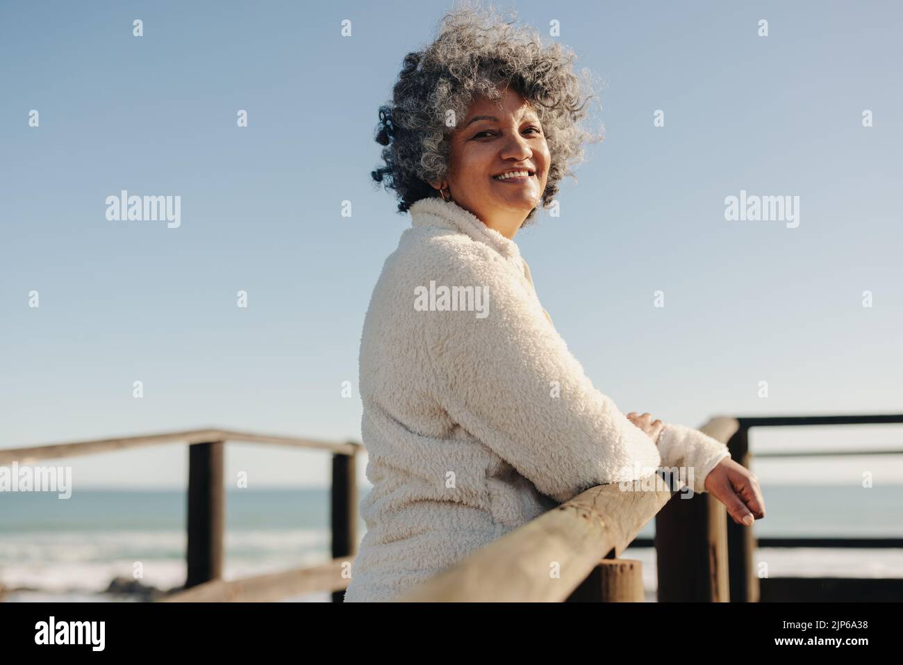 Bonne femme âgée souriant à la caméra tout en se tenant sur un pont de pied en bois à la plage. Joyeuse femme âgée appréciant des vacances rafraîchissantes à l'arrière Banque D'Images