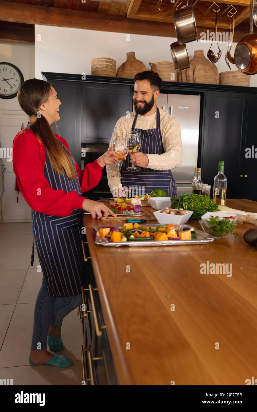 Joyeux couple de caucasiens préparant la nourriture dans la cuisine, faisant un toast avec des verres de vin blanc Banque D'Images