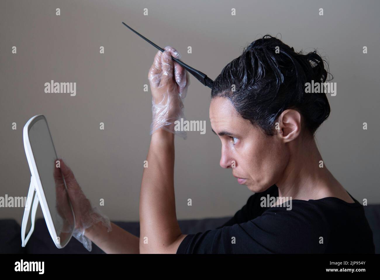Femme d'âge moyen colorant les cheveux foncés avec des racines grises à la maison. Femme assise sur un canapé et teinture des cheveux à l'aide d'une brosse noire et regardant le mirule Banque D'Images