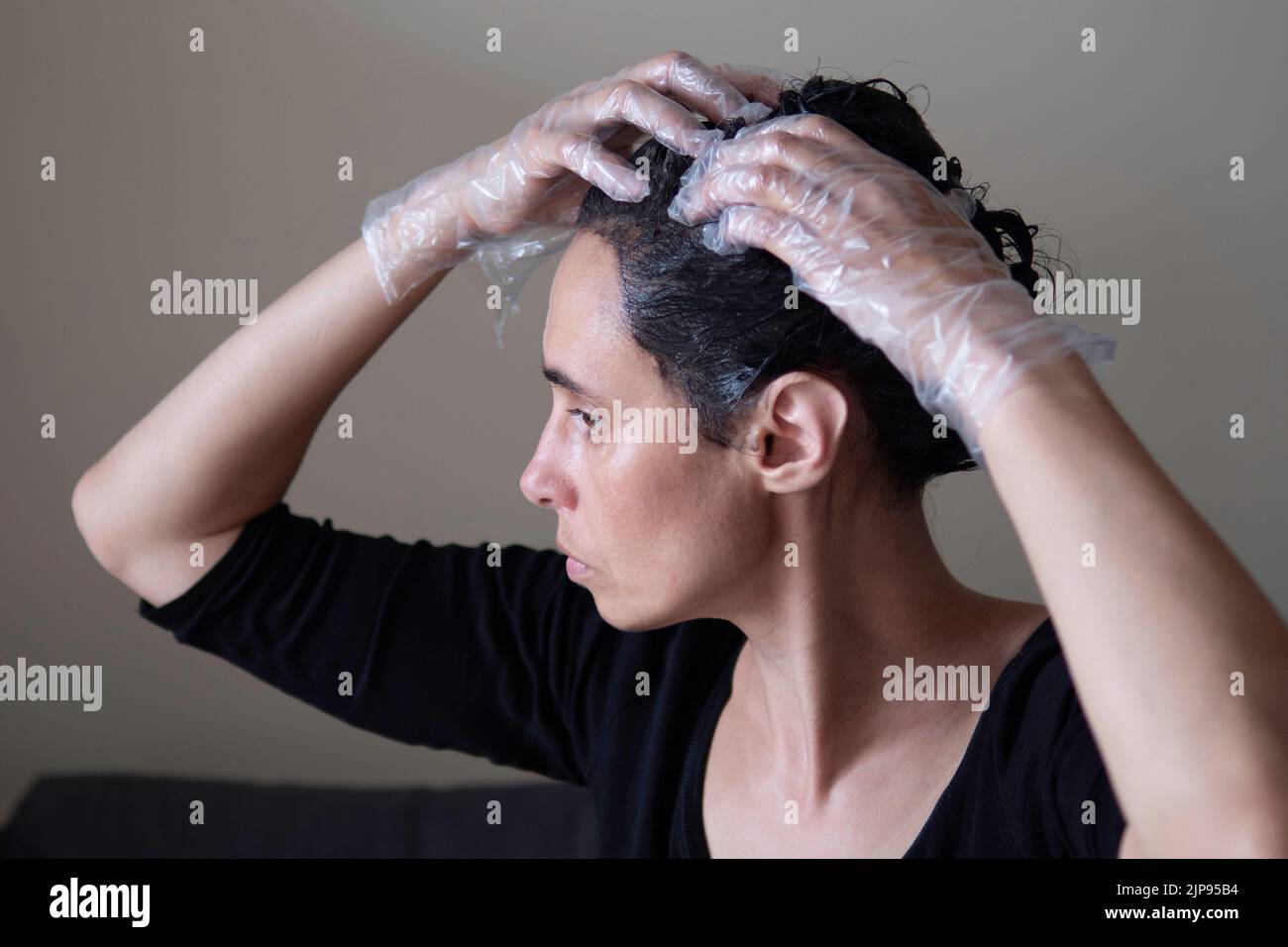 Femme d'âge moyen colorant les cheveux foncés avec des racines grises à la maison. Femme assise sur un canapé et teinture des cheveux à l'aide d'une brosse noire et regardant le mirule Banque D'Images