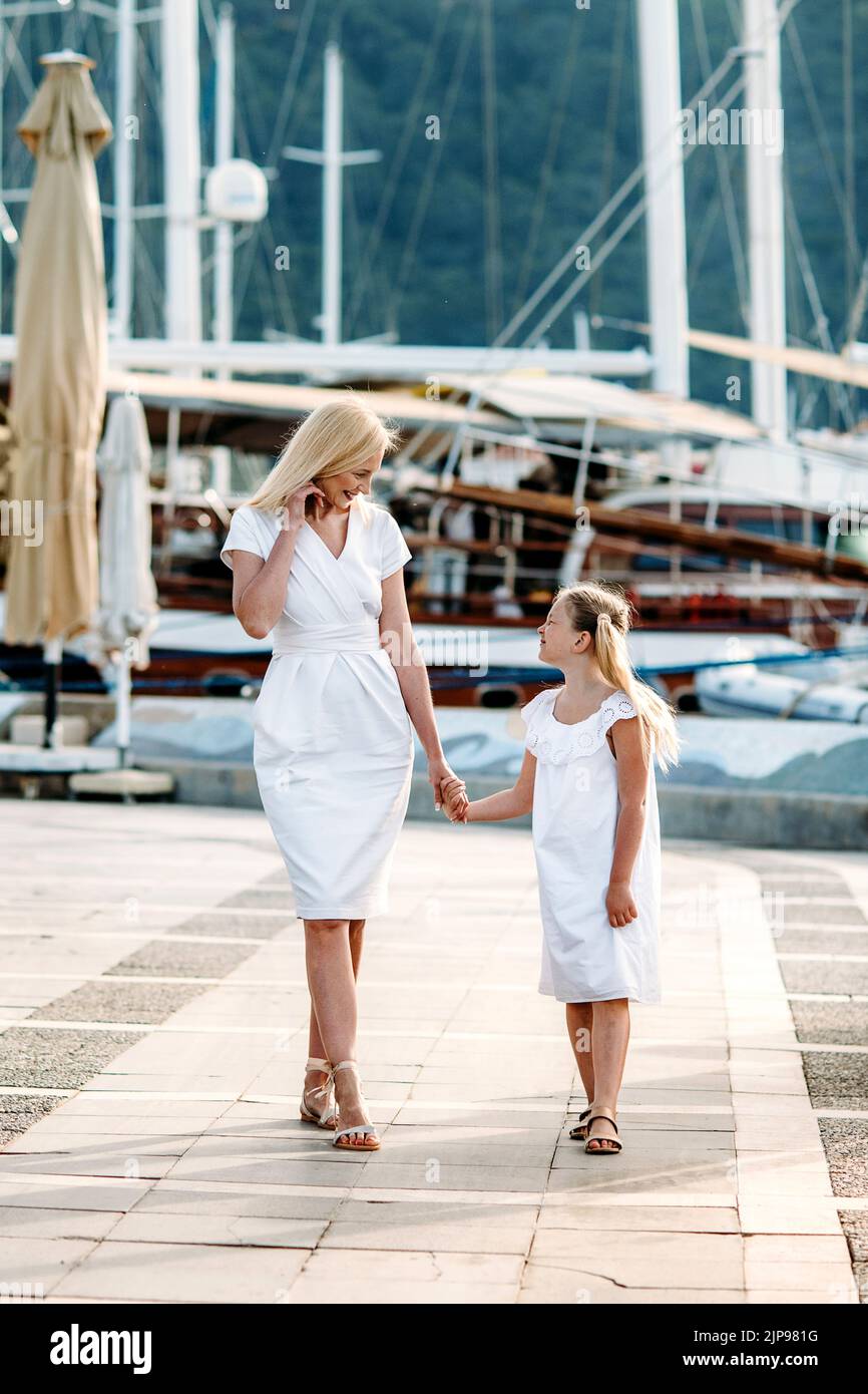 Bonne mère millénaire et fille blonde adolescente vêque blanche marchant près du port et de la marina avec des yachts dans une station balnéaire de luxe touristique wi Banque D'Images
