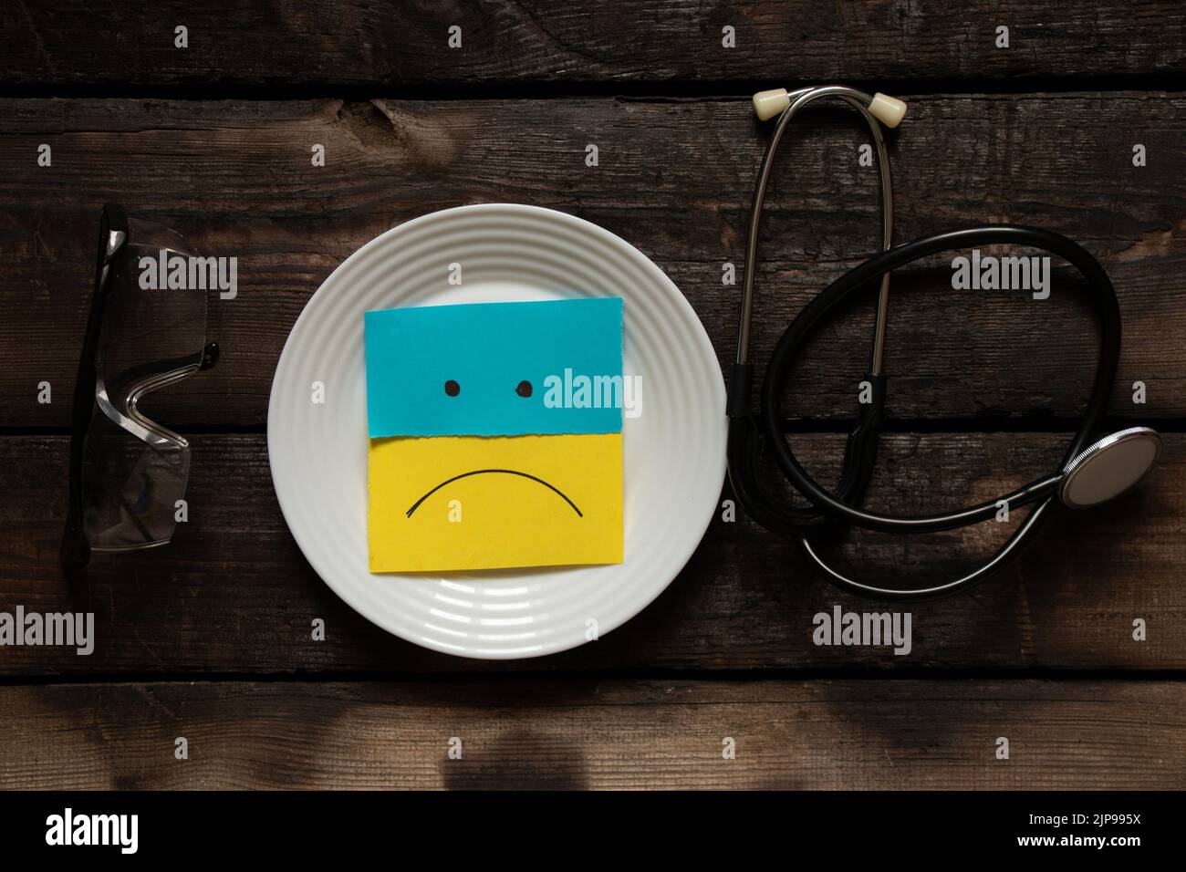 Le drapeau de l'Ukraine est jaune et bleu sur papier et un triste sourire peint dans une assiette sur la table. À proximité se trouve un stéthoscope et des lunettes médicales, arrêt t Banque D'Images
