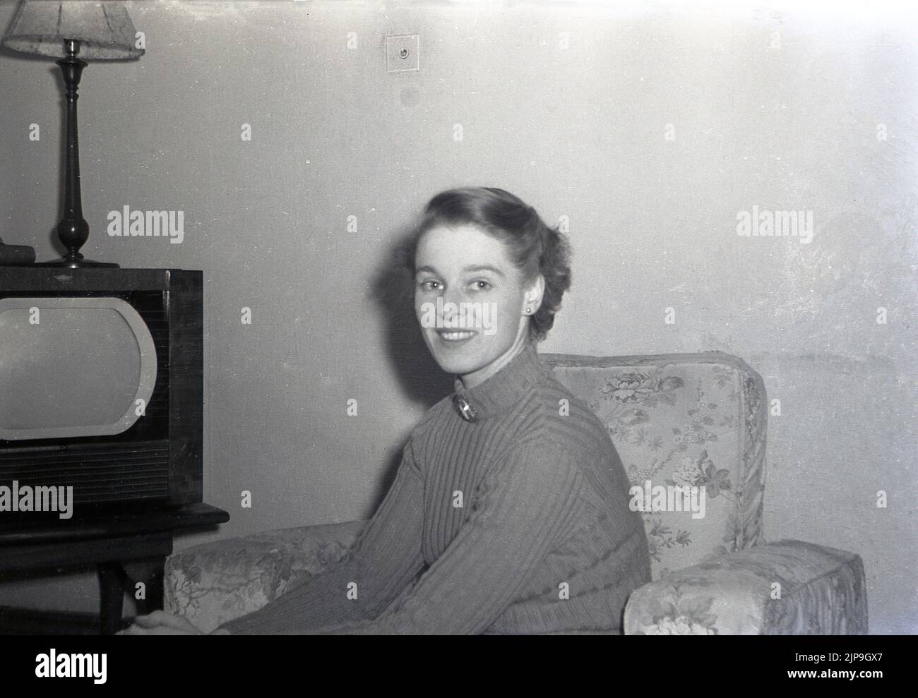 1953, historique, une jeune femme dans un chandail assis dans une chaise longue à côté d'un petit téléviseur de l'époque, Angleterre, Royaume-Uni. Comme de nombreux téléviseurs de l'époque, l'écran, de forme angulaire, était enfermé dans une armoire en bois. Banque D'Images